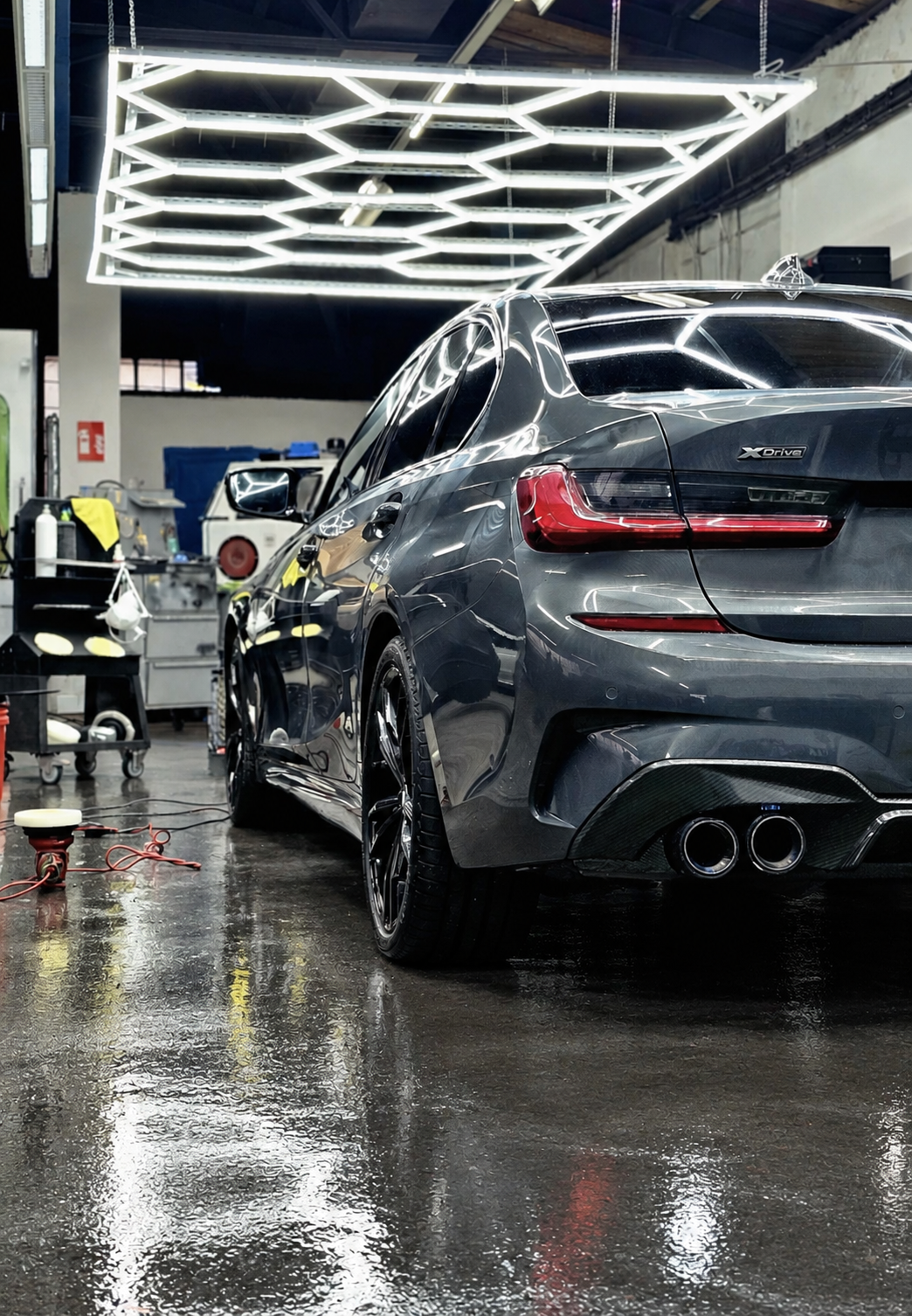 Dunkler BMW in Heckansicht unter der sechseckigen Werkstattbeleuchtung – Werkstattboden digital bearbeitet.